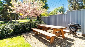 Une table de pique-nique en bois avec des bancs, située dans un jardin, avec un barbecue et un arbre aux fleurs roses.