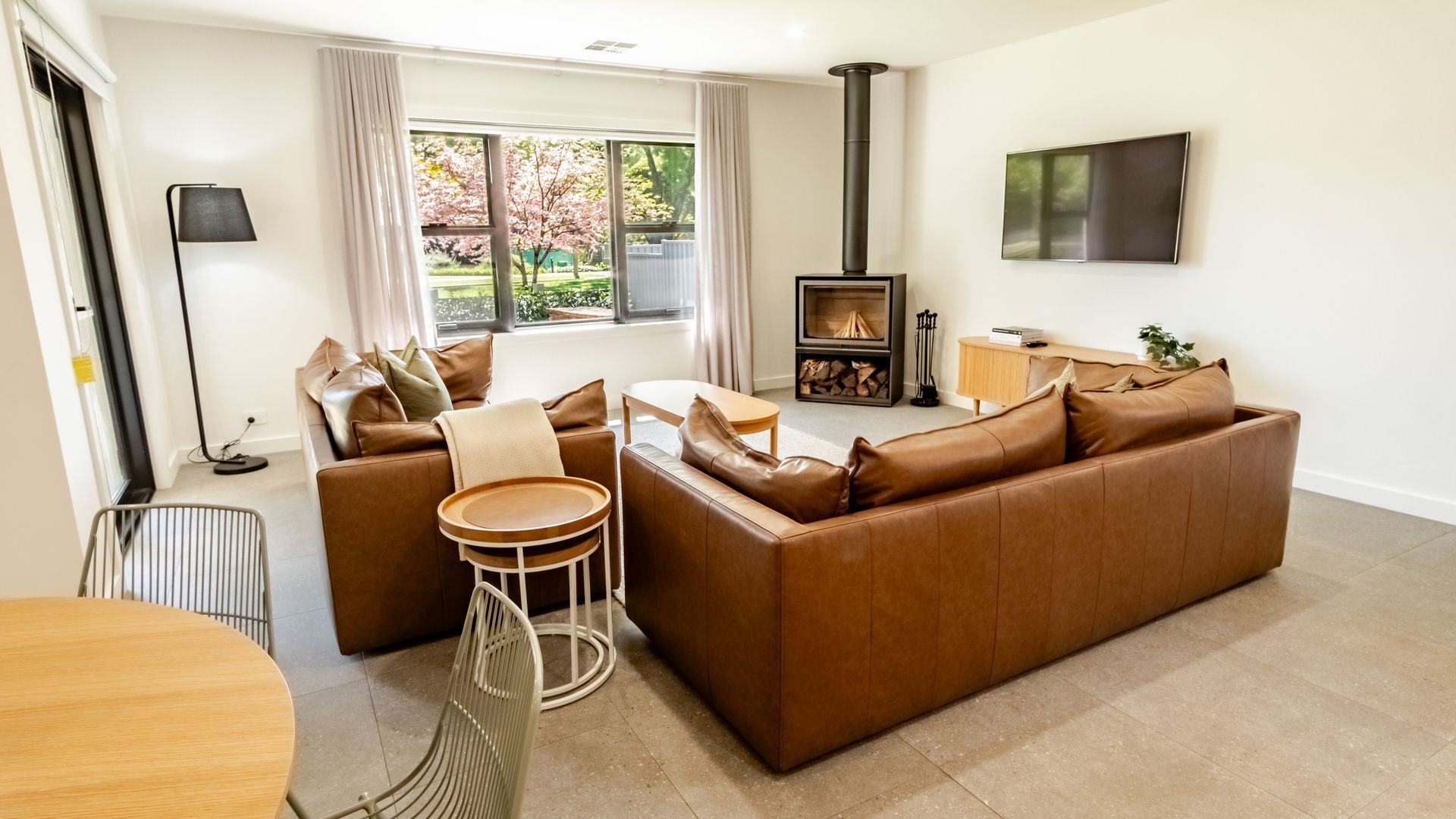 A modern living room with a brown leather sofa, a round wooden table, and a television mounted on the wall.
