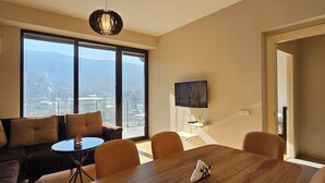 Una sala de estar moderna con una gran mesa de comedor, asientos cómodos, un televisor y vistas de un paisaje urbano a través de amplios ventanales.