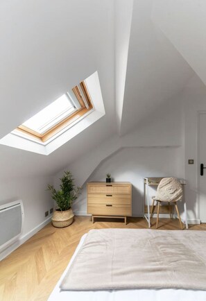 Ein Schlafzimmer mit Schrägdecke, einem Oberlicht, einem Bett, einer hölzernen Kommode, einem Stuhl und einer Topfpflanze.