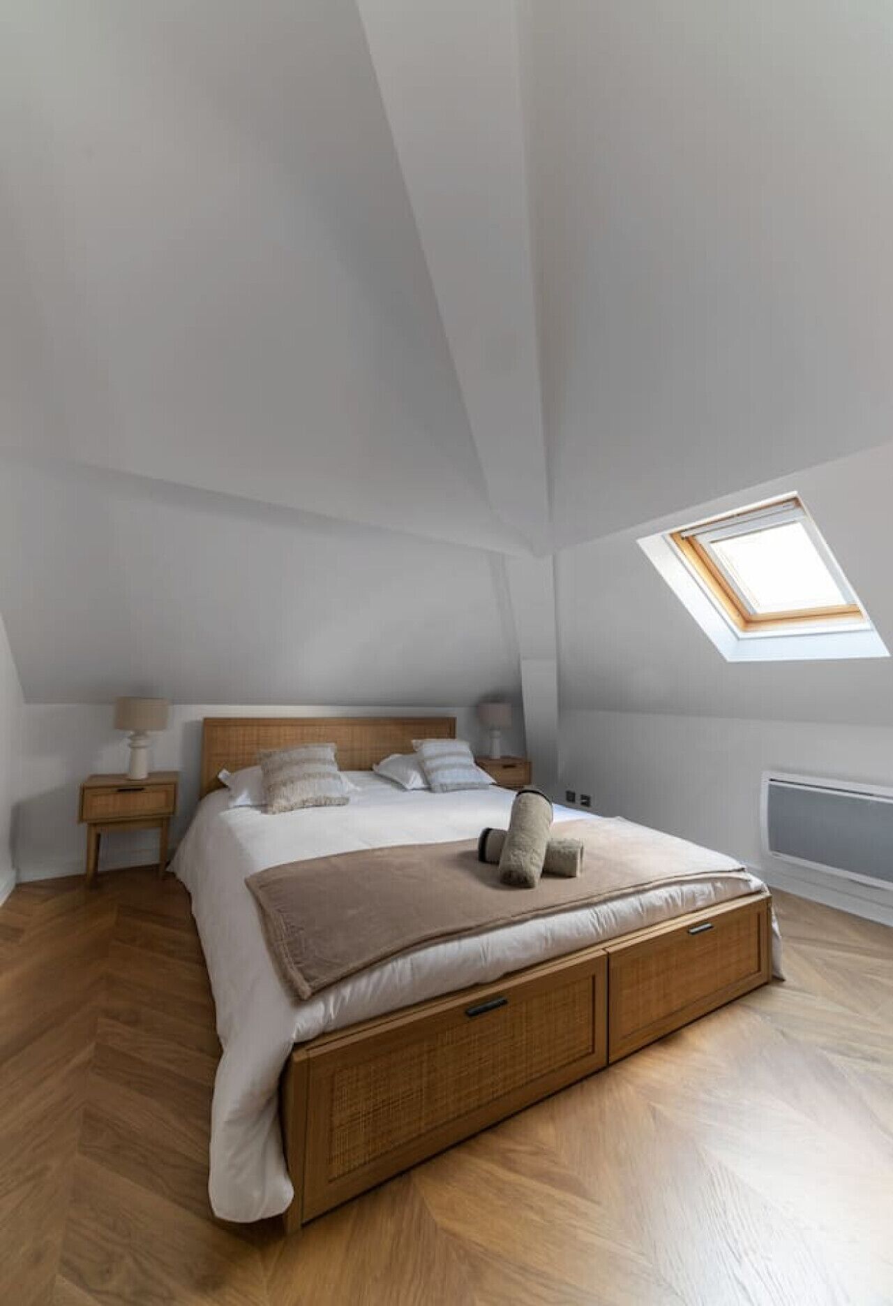 Een slaapkamer met een schuine kap, dakraam, bed met houten hoofdeinde, twee nachtkastjes met lampen en een radiator.