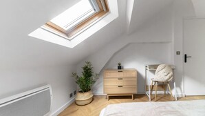 Een slaapkamer met een schuine kap, dakraam, een houten dressoir, een stoel en een potplant.