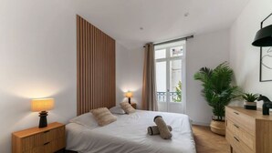 A modern bedroom with a large bed, wooden panel wall, bedside lamps, a potted plant, and a view of the outdoors.