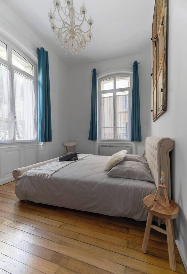 A bedroom with a bed, a chandelier, a window with curtains, and a wooden floor.