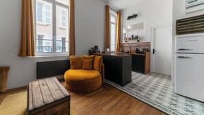 Ein gemütlicher, kompakter Wohnraum mit einer Couch, einem kleinen Holztisch, einer Küchenzeile mit Kühlschrank und einem Teppich mit Muster.