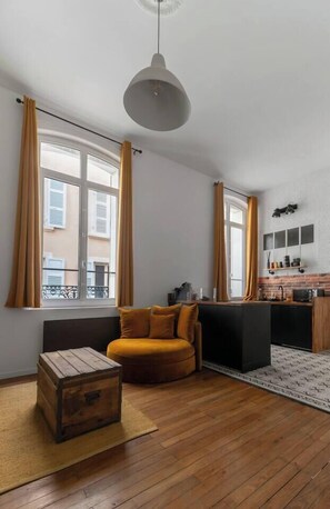 A modern living room with a wooden floor, a sofa, a small wooden chest, and a kitchen area with a brick backsplash and a hanging light fixture.