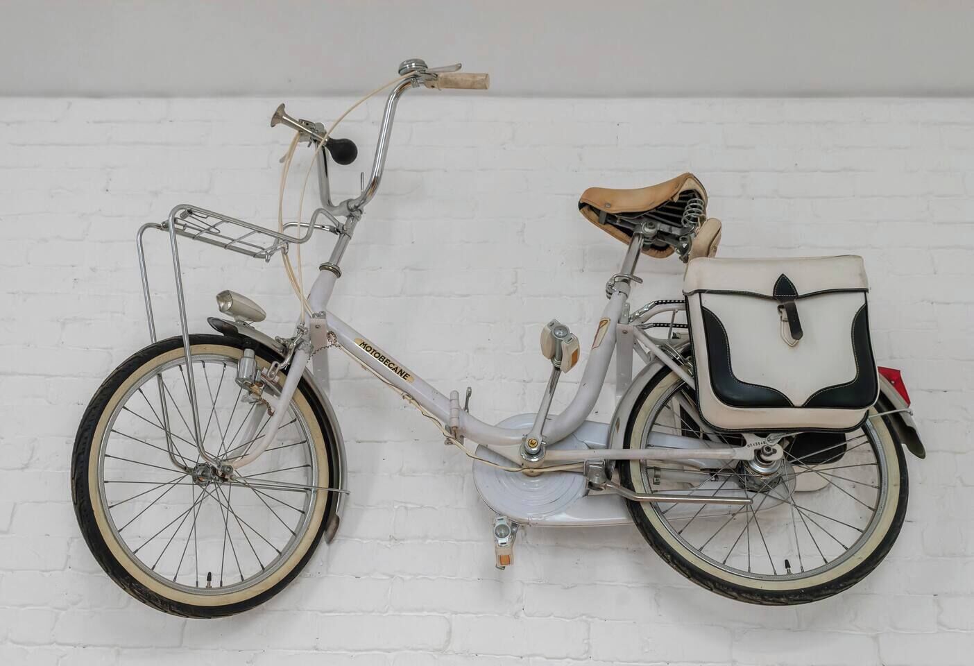 A vintage white bicycle with a leather seat and a front basket, leaning against a white brick wall.
