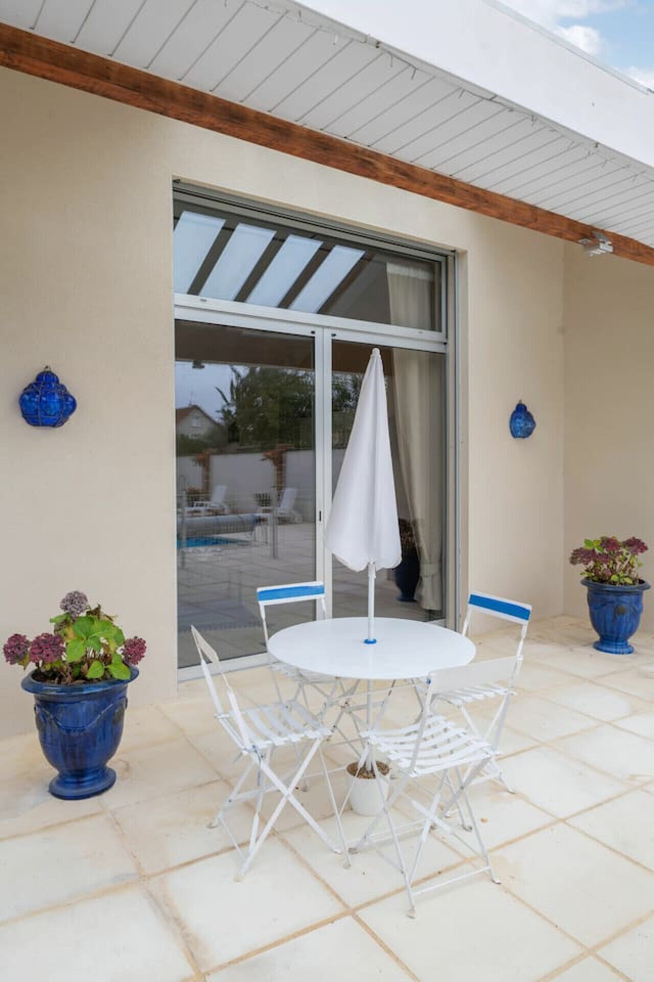 Un patio con una mesa y sillas blancas, dos plantas en macetas y un ventanal corredera.