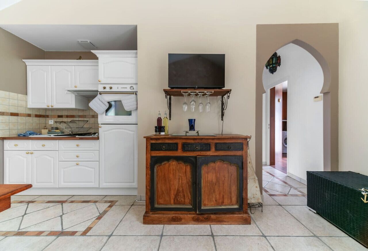 A kitchen with white cabinets, a wooden cabinet with a television above, and a tiled floor.