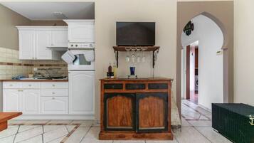 A kitchen with white cabinets, a wooden cabinet with a television above, and a tiled floor.