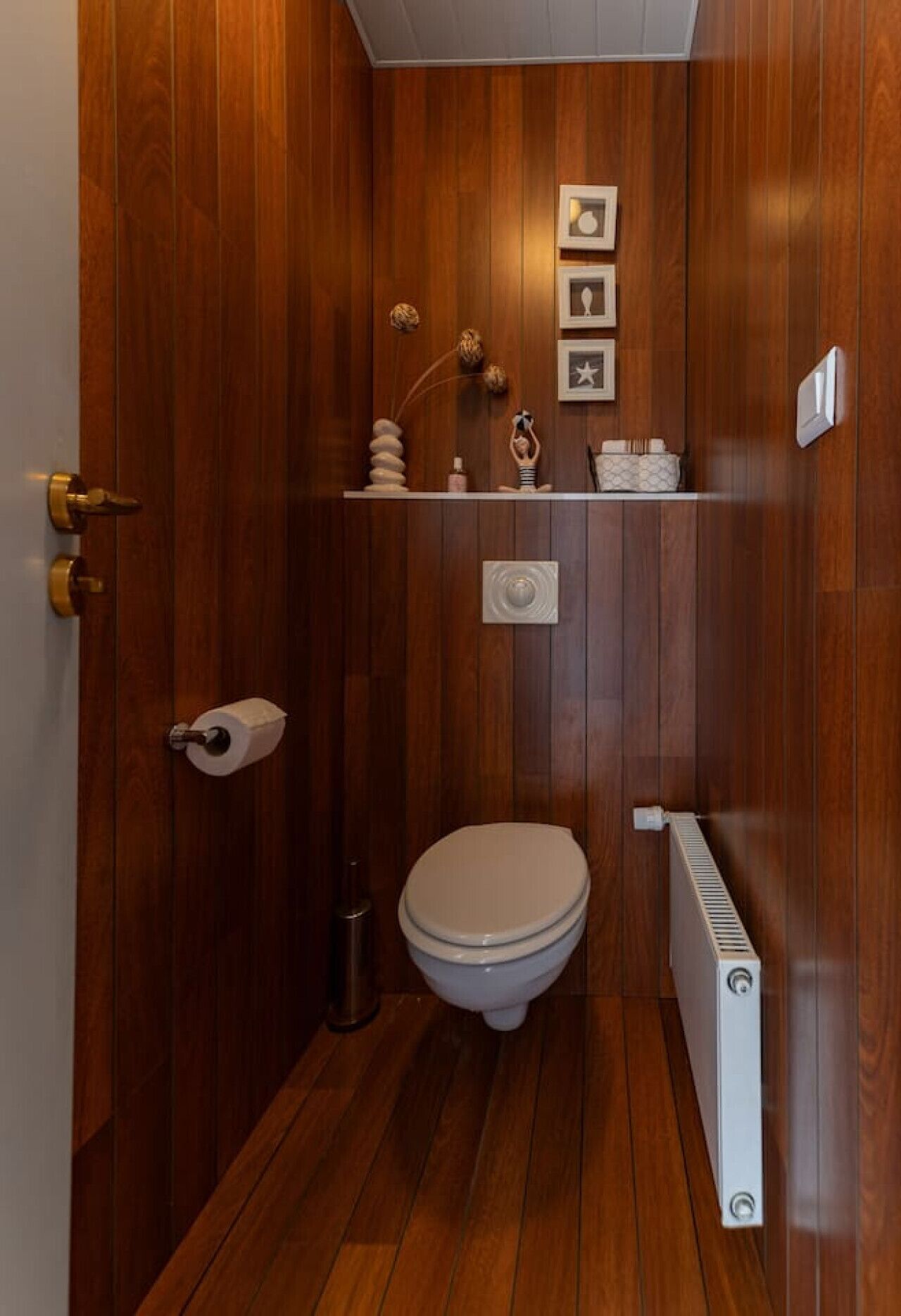 A compact wooden bathroom with a toilet, a radiator, and a small shelf with decorative items.