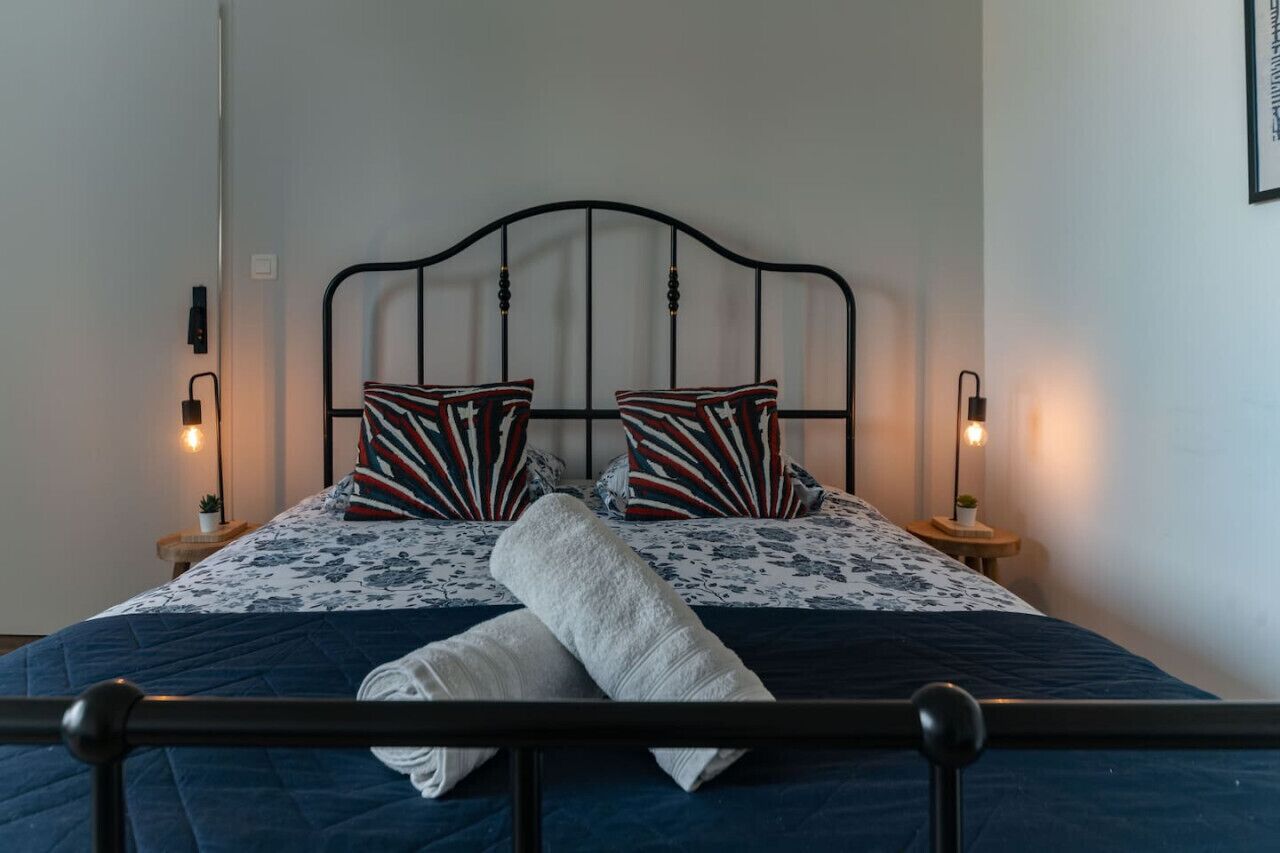 A neatly made bed with a blue quilt, two patterned pillows, and a white towel. There are bedside lamps on either side of the bed.
