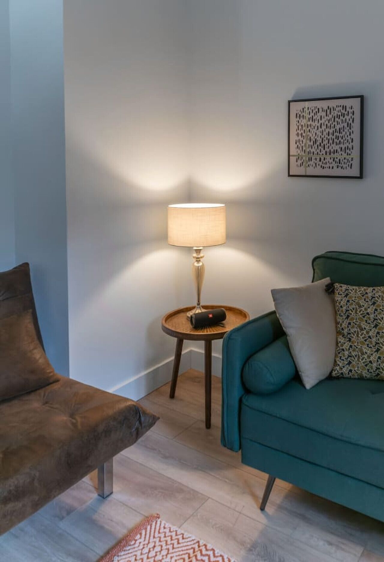 Un salon avec un fauteuil marron, un canapé turquoise, une petite table d’appoint en bois sur laquelle se trouve une lampe, et un coussin décoratif à motifs.