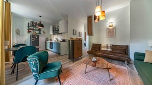 A modern living room with a sofa, coffee table, and chairs. There is a kitchen area with a dining table and chairs. The room has a large window and a hanging light fixture.