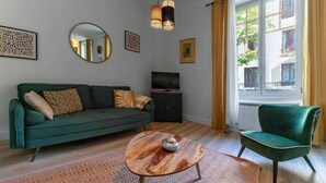 A living room with a green sofa, a round coffee table, a green armchair, a TV on a cabinet, and a window with curtains.