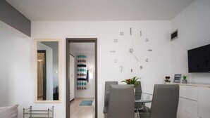 A modern living room with a sofa, a dining area with chairs and a table, a large wall clock, and a TV mounted on the wall. - Beautiful Apartment With Parking Space (Porec)