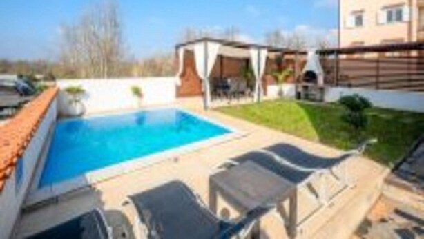 A swimming pool with lounge chairs, a barbecue area, and a building in the background. - Beautiful Apartment With Parking Space (Porec)