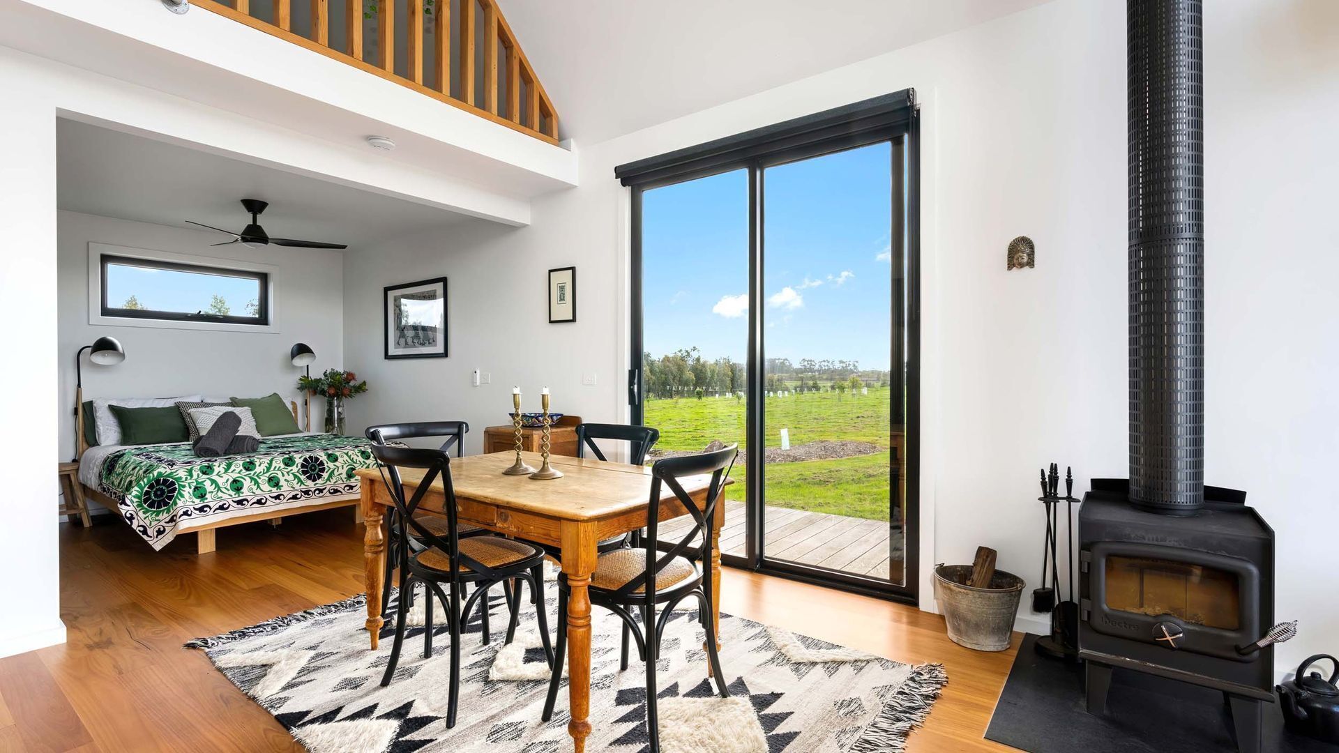 A modern living room with a dining area, a large window with a view of a green field, and a wood-burning stove.