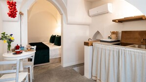 Una habitación compacta con cama, una mesita con artículos de desayuno, una cesta de fruta colgante y una zona de cocina con lavamanos y encimera.