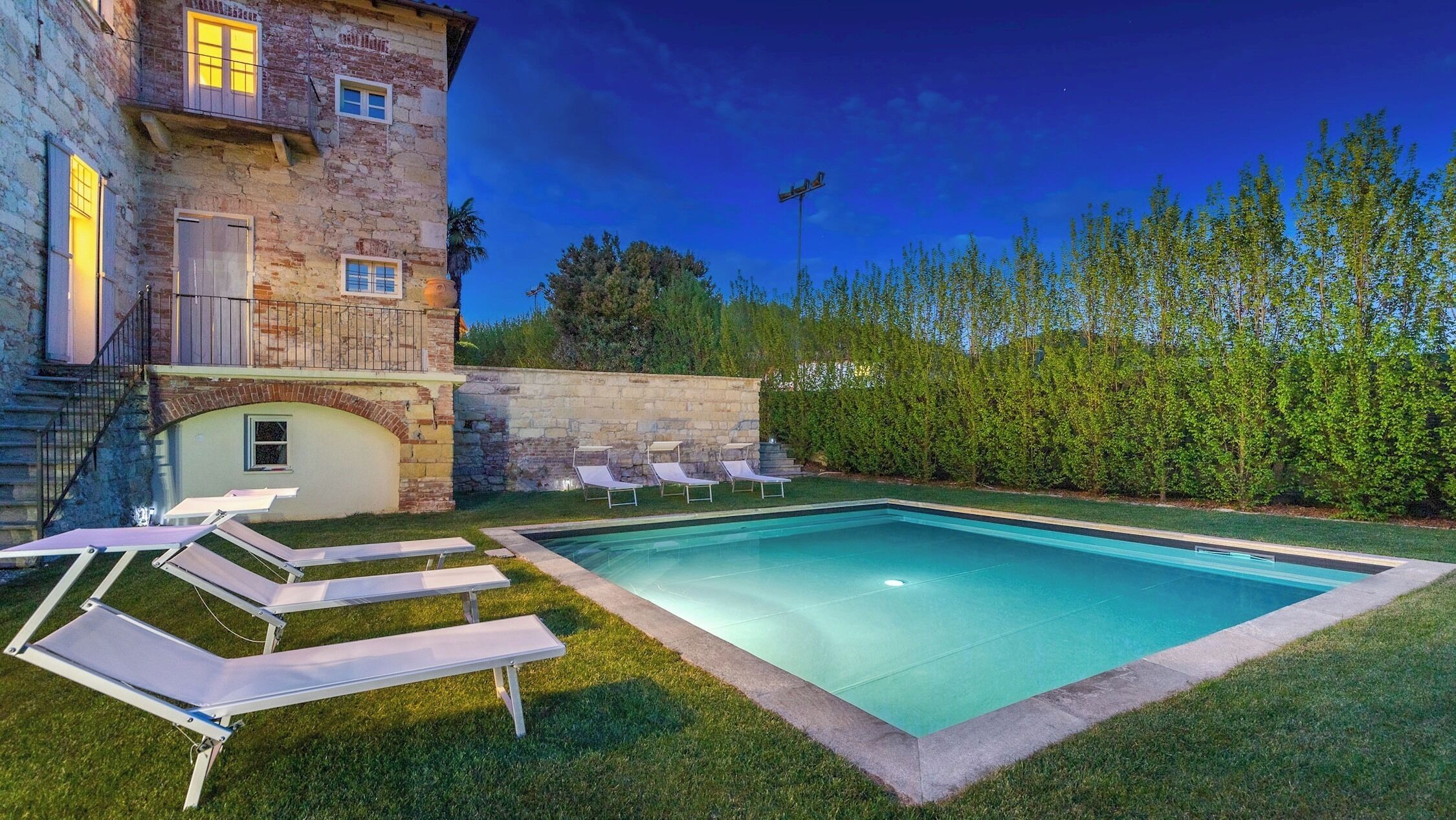 Une piscine avec des chaises longues, un bâtiment en pierre doté d’un balcon et une haie en arrière-plan.