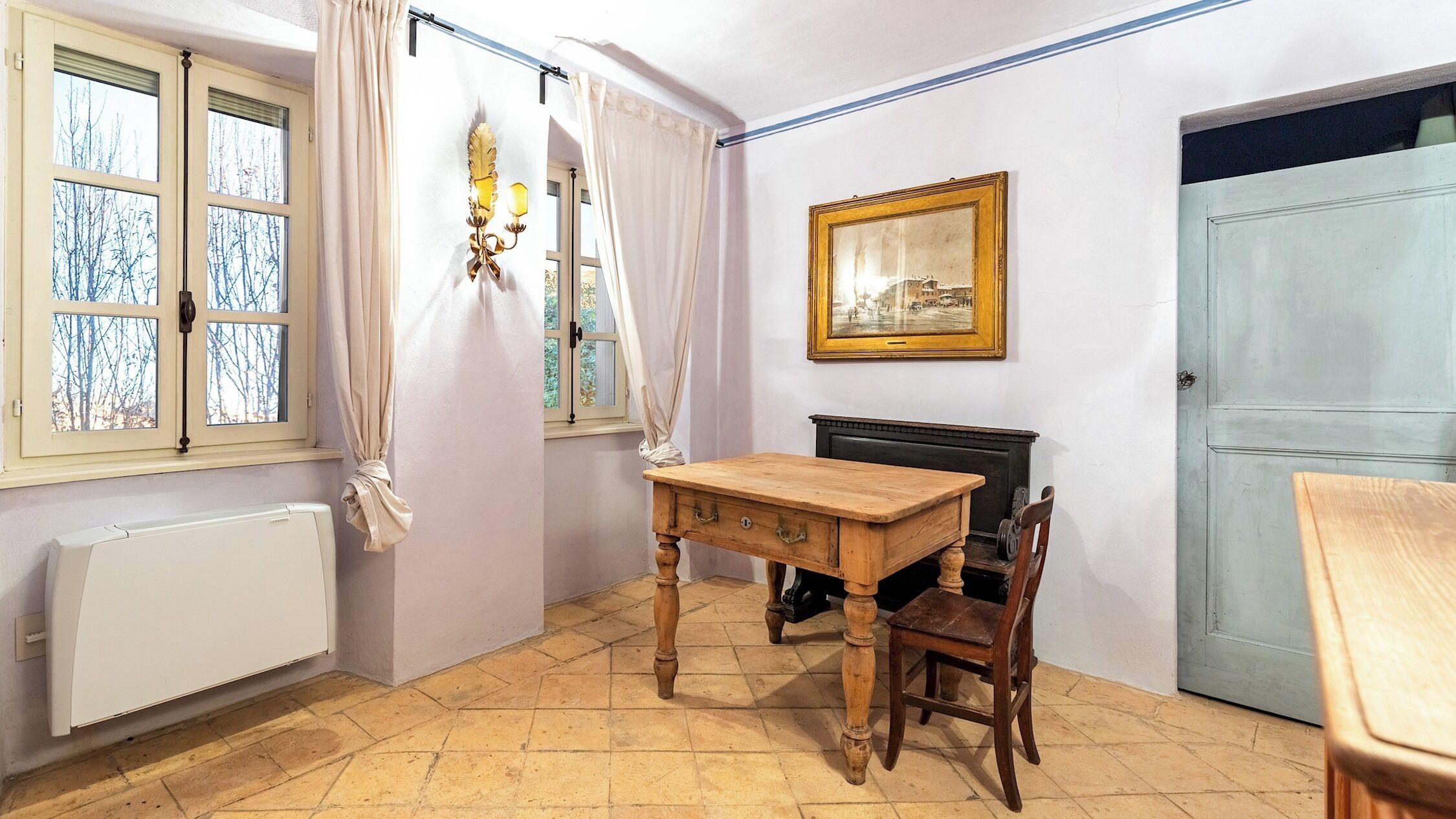 Una habitación con una mesa de madera, una silla, un radiador, una ventana con cortinas, un cuadro en la pared y una puerta.