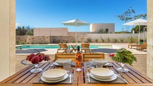 Une table à manger dressée avec des assiettes, des verres et une bouteille, donnant sur une piscine et un espace de détente extérieur.