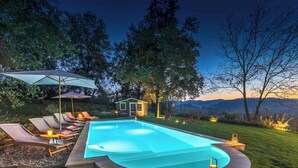 A swimming pool with lounge chairs and umbrellas, surrounded by trees and a small house in the background. - Cascina Monferrato 12, Emma Villas (Sala Monferrato)