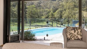 Un balcon donnant sur une piscine, entouré d’une rambarde en verre et bordé d’un paysage verdoyant luxuriant.