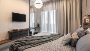 A modern bedroom with a large bed, a TV mounted on the wall, a bedside table, and a large window with curtains.