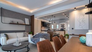 A modern living room with a sofa, coffee table, and dining table. There is a kitchen in the background with a TV and cabinets.