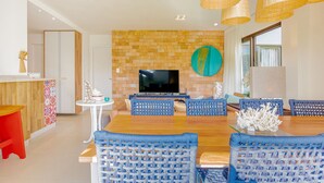 Un comedor moderno con una mesa de madera y sillas tejidas azules, un televisor montado en una pared de ladrillo y lámparas colgantes de mimbre.
