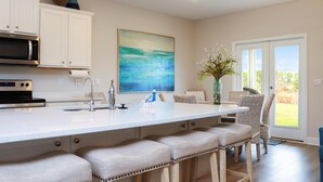A kitchen with white cabinets, a large island with bar stools, a stainless steel microwave, and a painting on the wall. - Coastal Oasis (Port St. Joe)