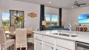 A modern kitchen with a white countertop, stainless steel sink, and white cabinetry. There is a dining area with wicker chairs and a wooden table. A television is mounted on the wall, and a ceiling fan is visible. - Coastal Oasis (Port St. Joe)