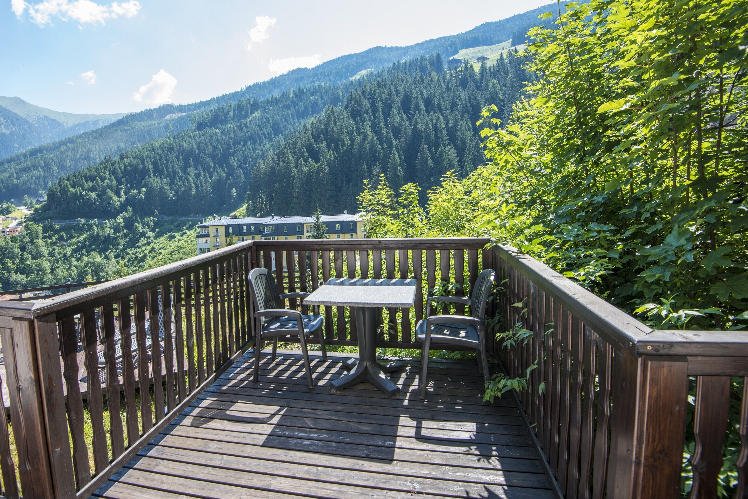 Een houten terras met een tafel en stoelen, met uitzicht op een bebost dal en bergen.