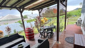 A covered outdoor seating area with a table and chairs, a red barrel, and a view of a house and greenery.