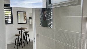 A modern bathroom with a shower, a wooden bench with a plant, and a view of a dining area with stools and a table.