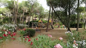 A playground with a wooden climbing structure and slides, surrounded by flowering plants and trees.