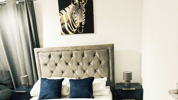 A bedroom with a tufted headboard, a bed with white and dark blue bedding, a zebra artwork on the wall, and a nightstand with a lamp. - Convenient Home - Sleeps 6 - Parking - Garden (Northampton)