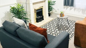 A modern living room with a fireplace, a flat-screen TV mounted above it, a grey sofa with cushions, and a patterned rug. - Convenient Home - Sleeps 6 - Parking - Garden (Northampton)