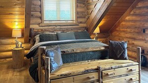 A log cabin bedroom with a loft bed, wooden walls, a window with blinds, and a small bedside table with a lamp.