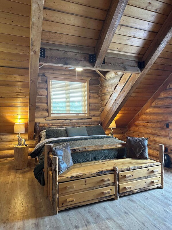 Un dormitorio de cabaña de madera con cama en altillo, paredes de madera, una ventana con persianas y una mesita de noche con lámpara.