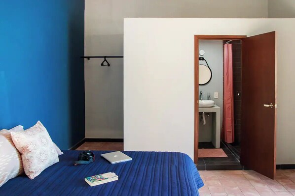 Een slaapkamer met een blauwe spreideken, een laptop en een camera op het bed. Een deur leidt naar een badkamer met een wastafel en douche.