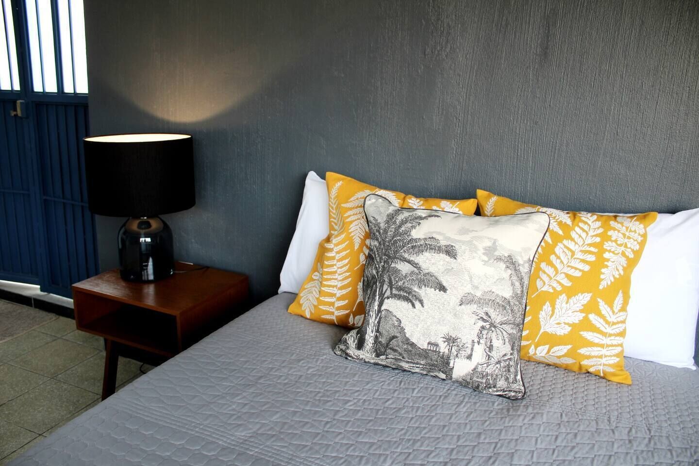 A neatly made bed with a grey quilt, a bedside table with a lamp, and two pillows with leaf patterns.