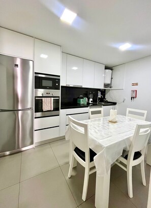 A modern kitchen with white cabinetry, a stainless steel refrigerator, a black countertop, and a dining table with white chairs. - Casa Augusta 6 (Braga)