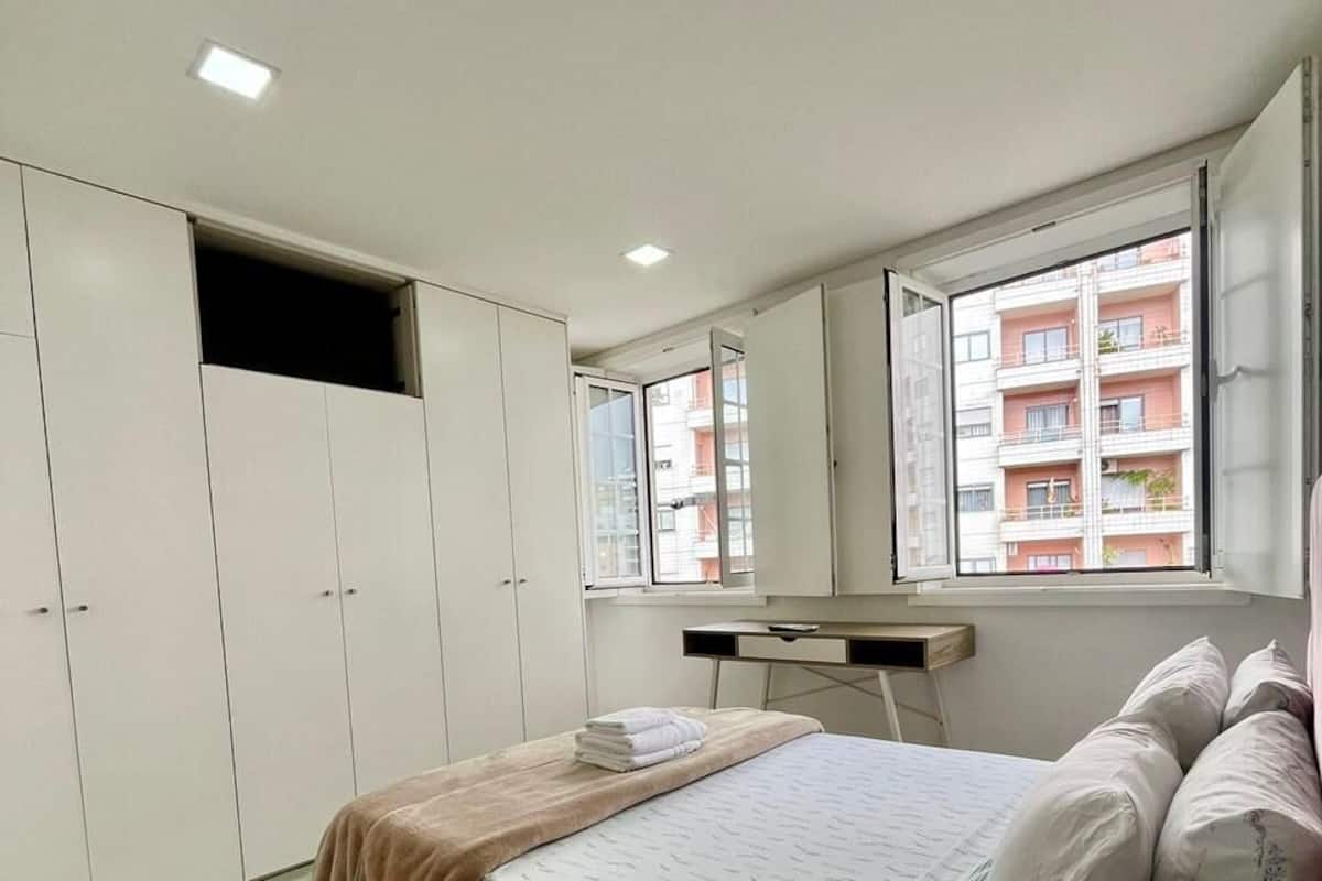 A bedroom with a bed, a desk, a wardrobe, and a window with a view of a building.