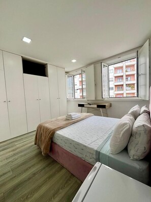 Ein Schlafzimmer mit Bett, Schreibtisch, Kleiderschrank und einem Fenster mit Blick auf ein Gebäude.