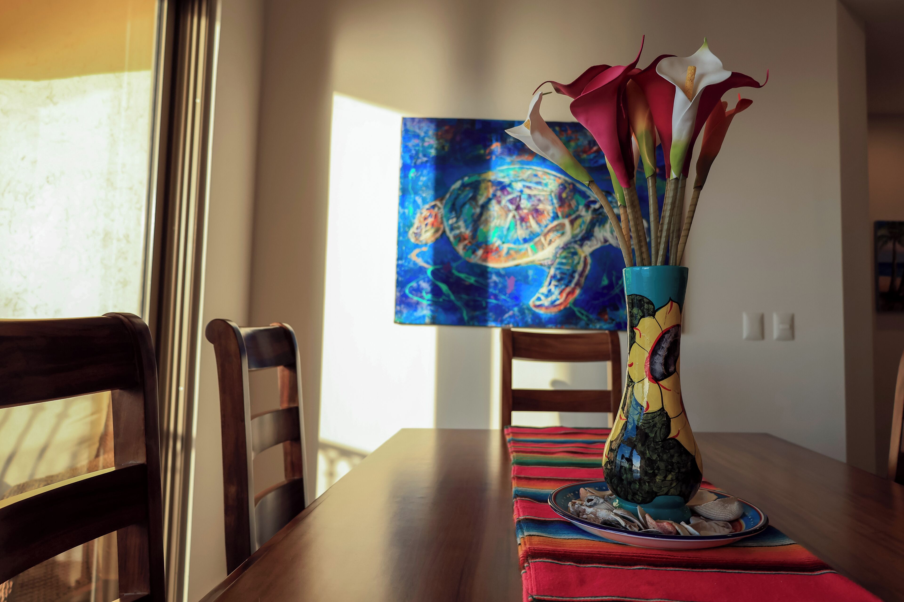 A dining table with a colorful vase of flowers, a plate with food, and a painting on the wall.