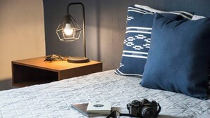 Een net opgemaakt bed met een gewatteerde spreideken, een camera en een paar schoenen. Er staat een nachtkastje met een lamp en een boekje. De kamer heeft kussens met een blauw-wit patroon.