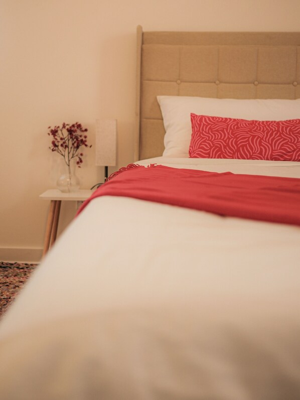 Una cama bien tendida con un edredón rojo y una almohada estampada, una mesita de noche con un florero y una lámpara.