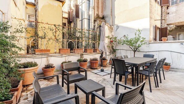 Un espace extérieur aménagé en terrasse, avec une table et des chaises, entouré de plantes en pot et bordé d’un bâtiment recouvert de lierre.